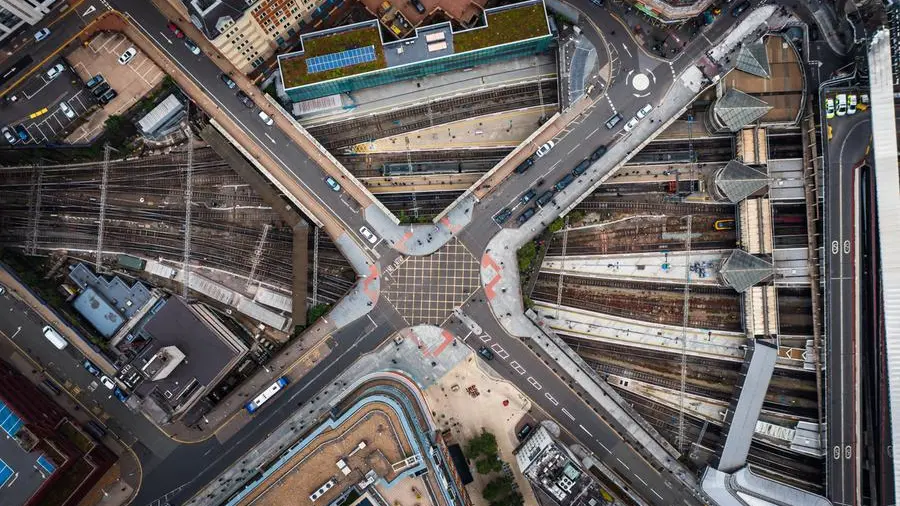 Piazza urbana con aree verdi e strade pavimentate, con persone che passeggiano