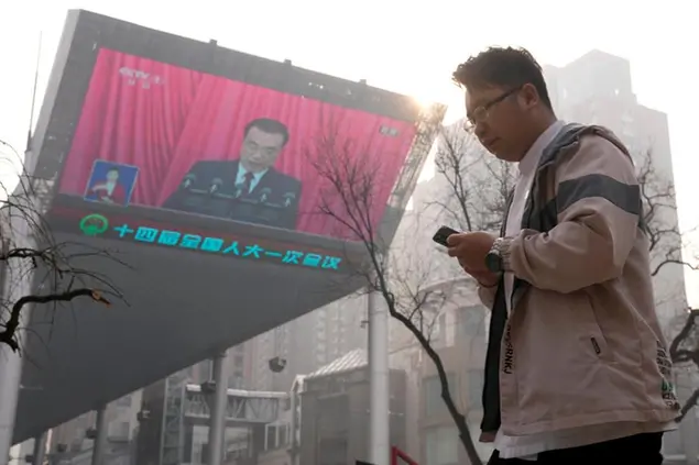 Un uomo cammina mentre sullo schermo si vede il premier uscente, Li Keqiang, che tiene il suo discorso\\u00A0(AP Photo/Andy Wong)