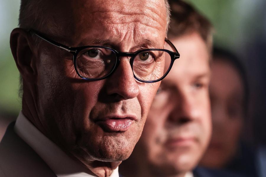 Un leader politico tedesco discute con un gruppo di persone in un contesto politico, con sfondo di un'immagine di un'area geografica