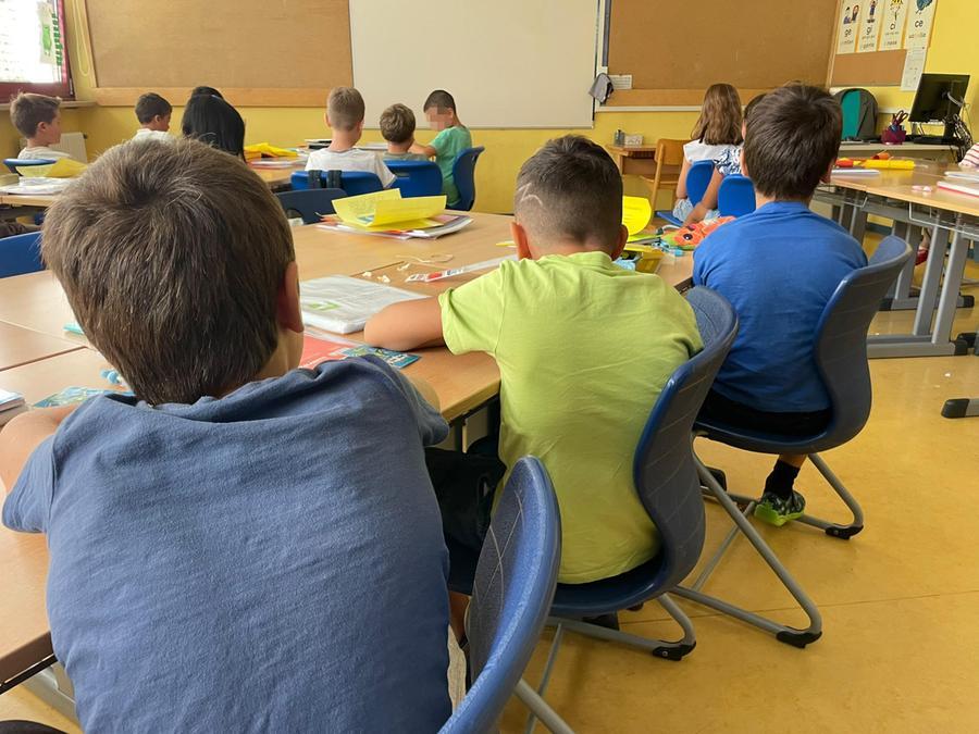 Un insegnante in classe con studenti, con un libro aperto e un'aula fresca, in un ambiente educativo che cerca di mantenere la distanza per un giudizio critico
