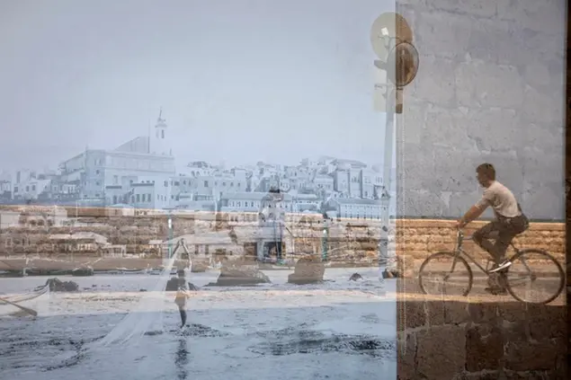 A man riding his bicycle is reflected on an old picture of Jaffa displayed in a window, in the Jaffa neighborhood of Tel Aviv, Israel, Wednesday, April 21, 2021. Historic Jaffa's rapid gentrification in recent years is coming at the expense of its mostly Arab lower class. With housing prices out of reach, discontent over the city’s rapid transformation into a bastion for Israel’s ultra-wealthy is reaching a boiling point. (AP Photo/Sebastian Scheiner)
