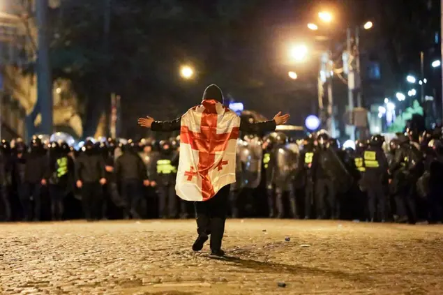 A protester wearing a Georgian national flag walks toward police line outside the Georgian parliament building in Tbilisi, Georgia, Tuesday, March 7, 2023. Georgian authorities used tear gas and water cannon outside the parliament building in the capital Tuesday against protesters who oppose a proposed law some see as stifling freedom of the press. (AP Photo/Zurab Tsertsvadze)