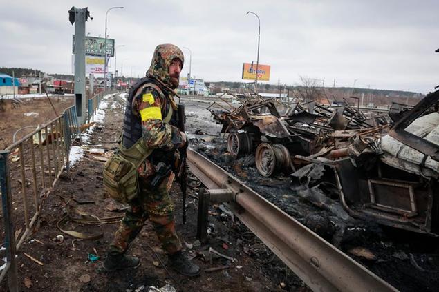 An armed man stands by the remains of a Russian military vehicle in Bucha, close to the capital Kyiv, Ukraine, Tuesday, March 1, 2022. (AP Photo/Serhii Nuzhnenko)