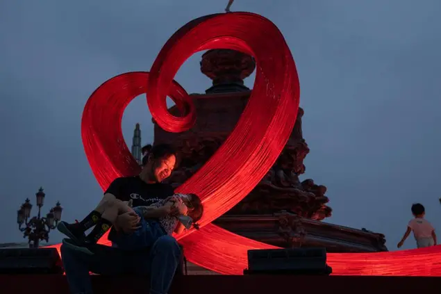A woman holds a child near heart shaped art installation around a fountain in Beijing on Aug. 14, 2021. China will now allow couples to legally have a third child as it seeks to hold off a demographic crisis that could threaten its hopes of increased prosperity and global influence. (AP Photo/Ng Han Guan)