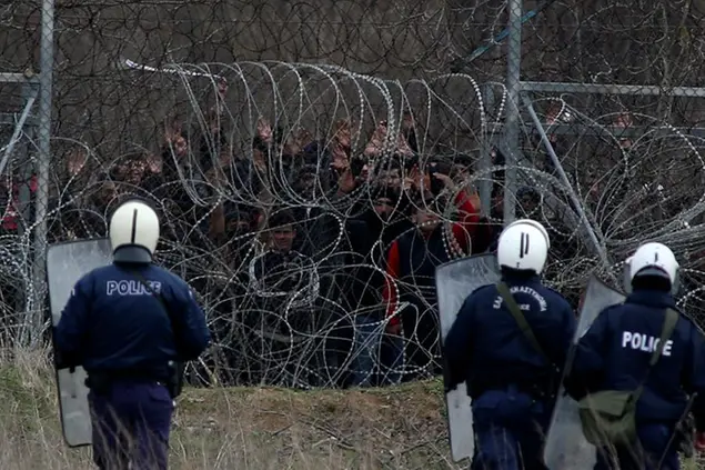 Greek riot police guard as migrants stand on a fence as they try to enter Greece from Turkey at the Greek-Turkish border in Kastanies on Wednesday, March 4, 2020. Facing a potential wave of nearly a million people fleeing fighting in northern Syria, Turkey has thrown open its borders with Greece to thousands of refugees and other migrants trying to enter Europe, and has threatened to send \\\"millions\\\" more. (AP Photo/Giannis Papanikos)