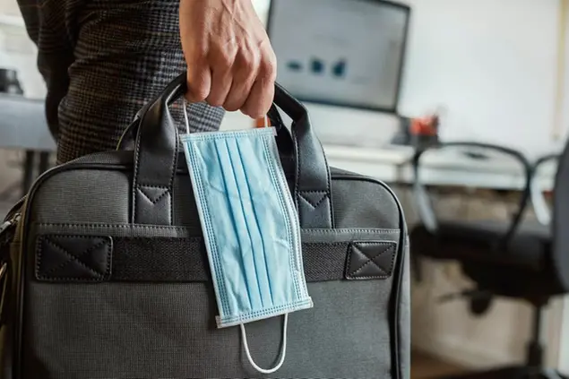 closeup of a young man in an office holding a briefcase and a surgical mask in his hand