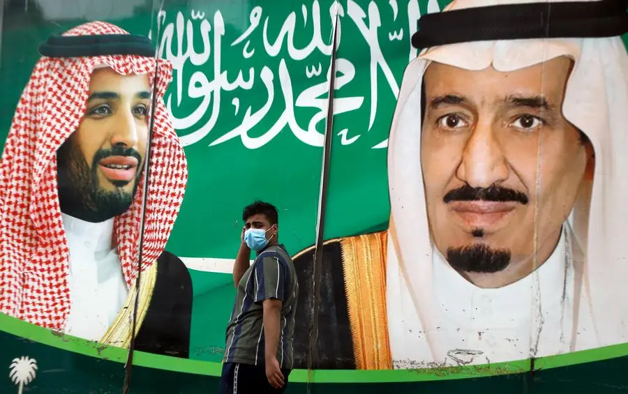 A man wearing a face mask to help curb the spread of the coronavirus walks past a banner showing Saudi King Salman, right, and his son Crown Prince Mohammed bin Salman, outside a mall in Jiddah, Saudi Arabia, Wednesday, Dec. 16, 2020. Saudi Arabia plans to cut more than 7% in spending next year, the finance ministry said, as the region's biggest oil producer seeks to trim a gaping budget deficit amid the coronavirus pandemic and falling oil prices. (AP Photo/Amr Nabil)