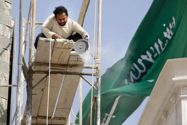 A Bengali worker cuts a brick while working on a mosque, as a giant Saudi flag billows in the background, at King Abdullah Square, in Jiddah, Saudi Arabia, Thursday, Jan. 28, 2021. An Islamic creed reads, \\\"there is no deity except God, and Muhmmad is the messenger of God.\\\" (AP Photo/Amr Nabil)